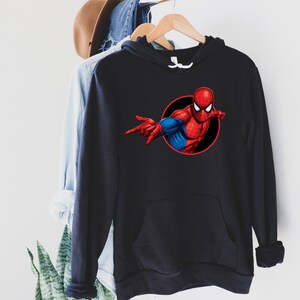 May include: Black hooded sweatshirt featuring a graphic of Spider-Man in a red and blue suit, set against a black circle. The superhero is depicted with his arm extended, pointing. The sweatshirt has a front pocket and a drawstring hood.