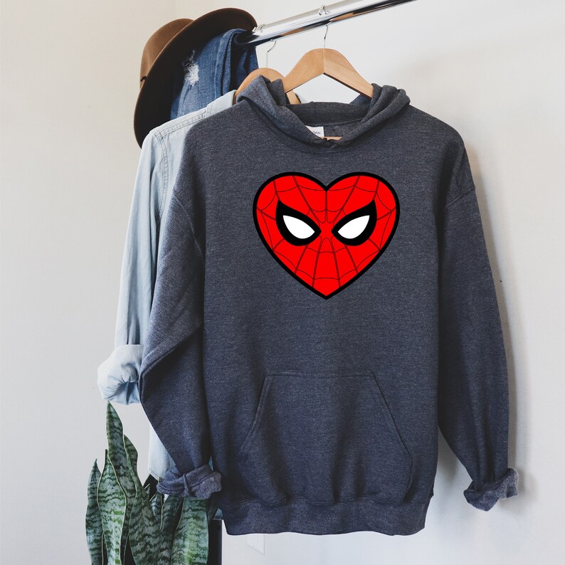May include: A dark gray hoodie with a red heart-shaped design featuring a spiderweb pattern and white eye shapes. The hoodie has a front pocket and is hanging on a wooden hanger.