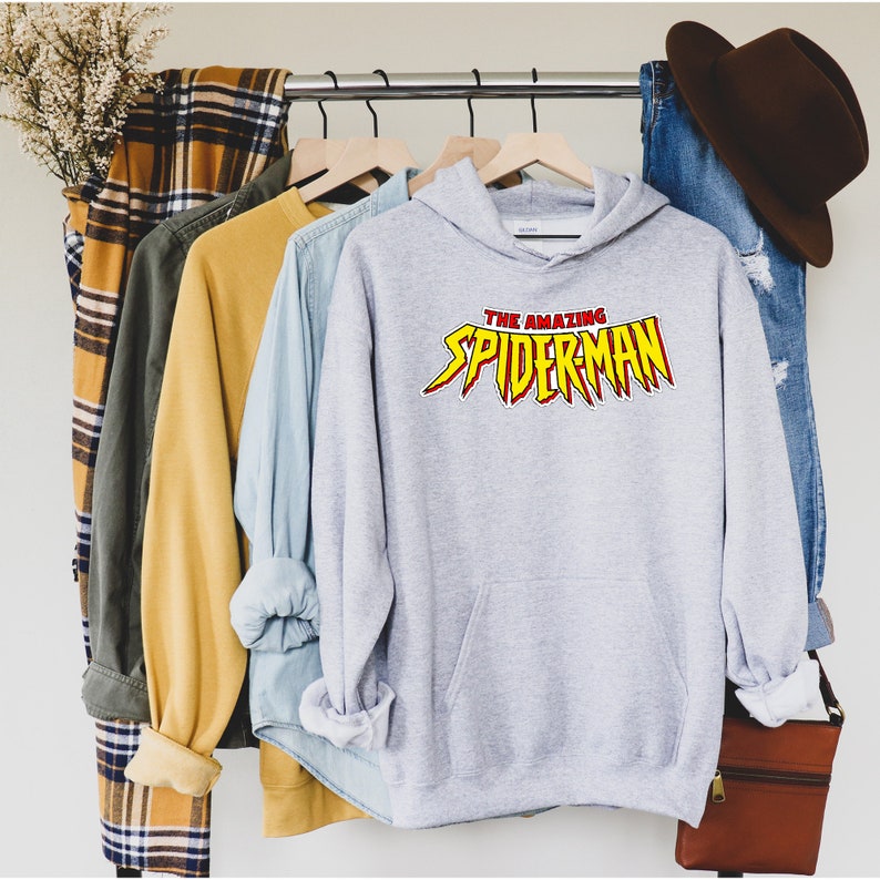 May include: A light gray hoodie with the text "THE AMAZING SPIDERMAN" in red and yellow. The hoodie is displayed on a clothing rack with other shirts and a pair of jeans.