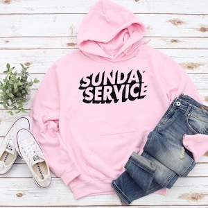 May include: A light pink hoodie with the text "SUNDAY SERVICE" in black, paired with blue jeans and white sneakers. The hoodie has a hood and a front pocket. The sneakers are white canvas. The jeans are folded at the bottom.