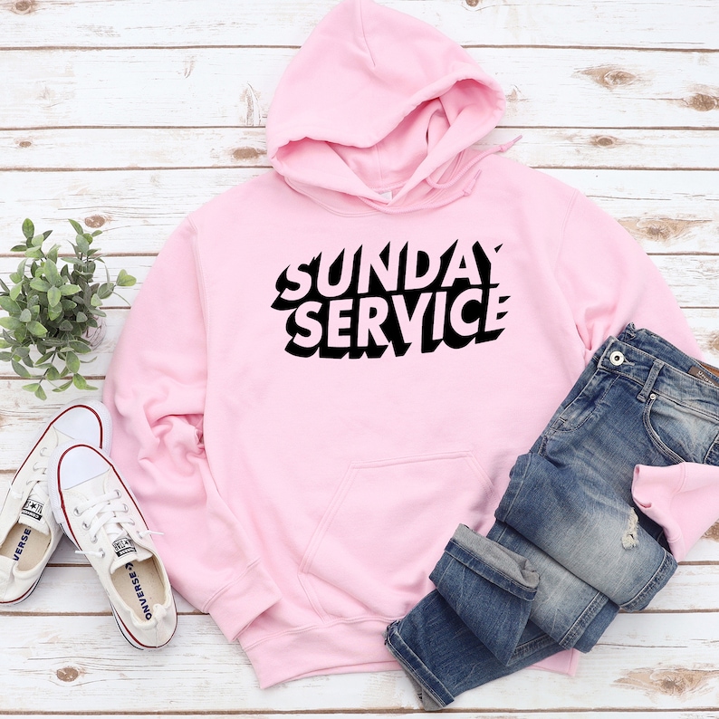 May include: A light pink hoodie with the text "SUNDAY SERVICE" in black, paired with blue jeans and white sneakers. The hoodie has a hood and a front pocket. The sneakers are white canvas. The jeans are folded at the bottom.