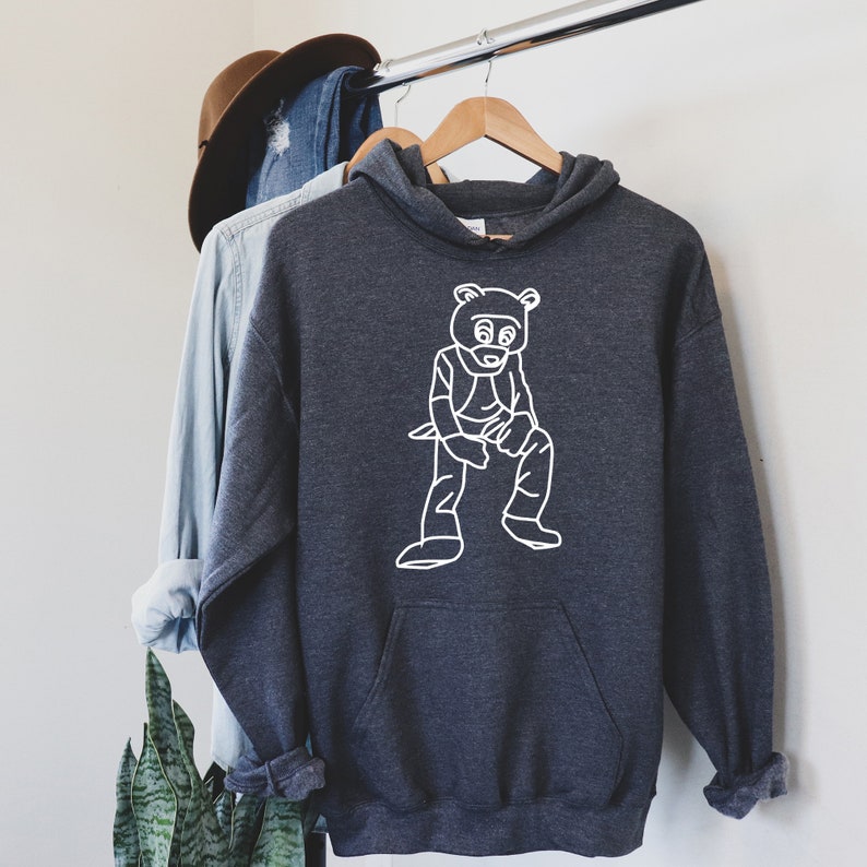 Pu&ograve; includere: Felpa con cappuccio grigio scuro con un'illustrazione bianca di un orso. La felpa ha una tasca a marsupio e un cappuccio con coulisse. L'indumento &egrave; appeso a una gruccia di legno.