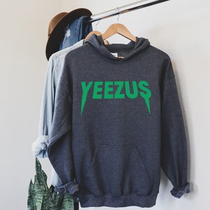 May include: A dark gray hoodie with the word "YEEZUS" printed in bright green across the chest. The hoodie has a hood and a front pocket. The text has a unique font with sharp, downward-pointing serifs. The hoodie is hanging on a wooden hanger.