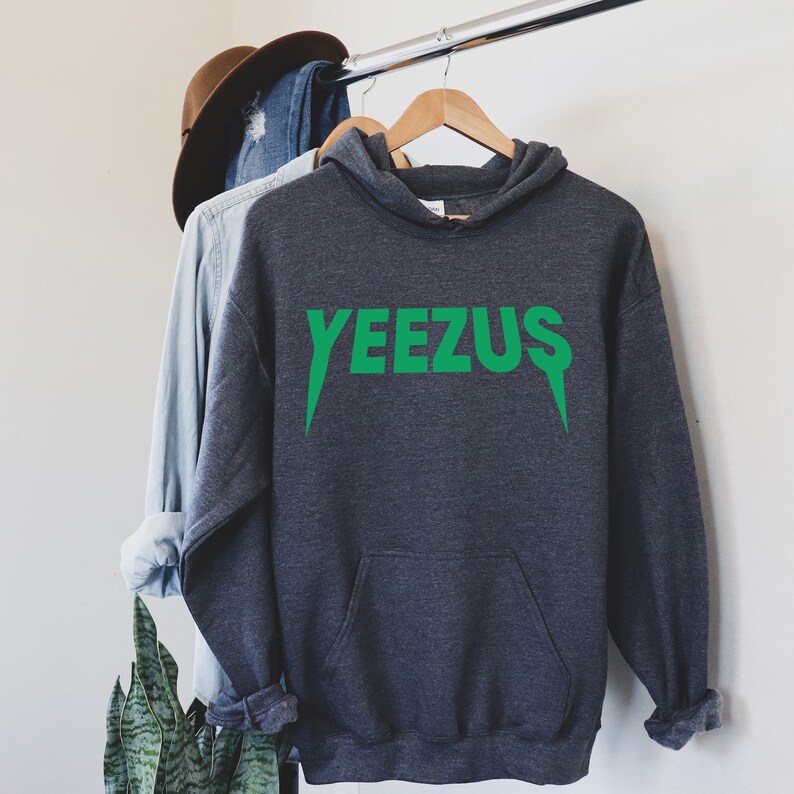 May include: A dark gray hoodie with the word "YEEZUS" printed in bright green across the chest. The hoodie has a hood and a front pocket. The text has a unique font with sharp, downward-pointing serifs. The hoodie is hanging on a wooden hanger.