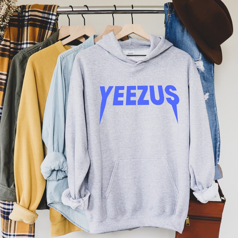 May include: A light gray hoodie with the word "YEEZUS" in bright blue lettering across the front. The hoodie has a front pocket and a hood. Other clothing items are visible on the rack, including a denim shirt, a yellow shirt, and a plaid shirt.