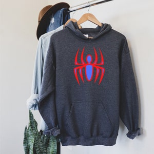K&ouml;nnte beinhalten: Dunkelgrauer Kapuzenpullover mit einem roten und blauen Spinnenmuster auf der Vorderseite. Der Hoodie hat eine K&auml;ngurutasche und eine Kapuze mit Kordelzug. Andere Kleidungsst&uuml;cke und ein Hut sind im Hintergrund zu sehen.