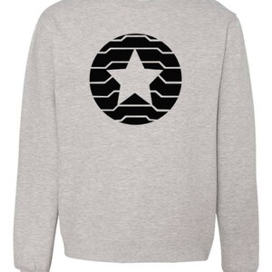 Puede incluir: Sudadera gris claro con un dise&ntilde;o de estrella negra dentro de un patr&oacute;n circular. La estrella est&aacute; centrada y el c&iacute;rculo est&aacute; compuesto por l&iacute;neas horizontales. La sudadera tiene cuello redondo y mangas largas.