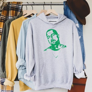 Op de afbeelding: Een lichtgrijze hoodie met een groene grafische afbeelding van een man. De hoodie heeft een voorzak en een capuchon. Andere kledingstukken zijn zichtbaar op een kledingrek, waaronder een geruit overhemd, een gele trui en een blauw overhemd met knopen.