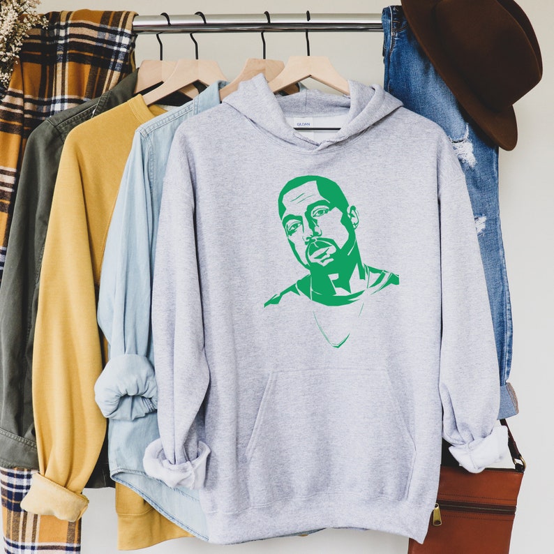 Op de afbeelding: Een lichtgrijze hoodie met een groene grafische afbeelding van een man. De hoodie heeft een voorzak en een capuchon. Andere kledingstukken zijn zichtbaar op een kledingrek, waaronder een geruit overhemd, een gele trui en een blauw overhemd met knopen.