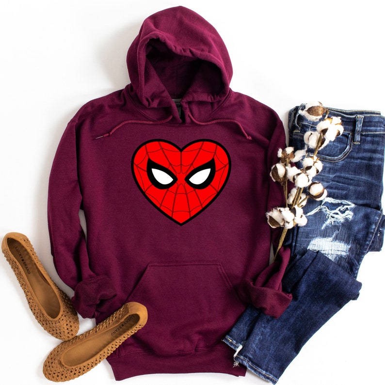 May include: Burgundy hooded sweatshirt featuring a heart-shaped Spider-Man mask design. The graphic is red with black outlines and white eye details. The image also shows blue jeans, brown flats, and cotton stems.