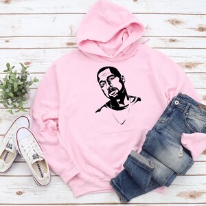 Op de afbeelding: Een lichtroze hoodie met een zwart-wit grafisch portret. De hoodie wordt gecombineerd met een blauwe spijkerbroek en witte canvas sneakers. De items zijn gerangschikt op een licht houtnerfoppervlak.