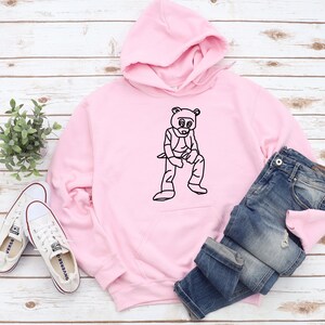 Pu&ograve; includere: Una felpa con cappuccio rosa chiaro con una grafica nera di una figura simile a un orso. La felpa &egrave; abbinata a jeans blu e scarpe da ginnastica di tela bianche. Gli articoli sono disposti su una superficie chiara con venature del legno.