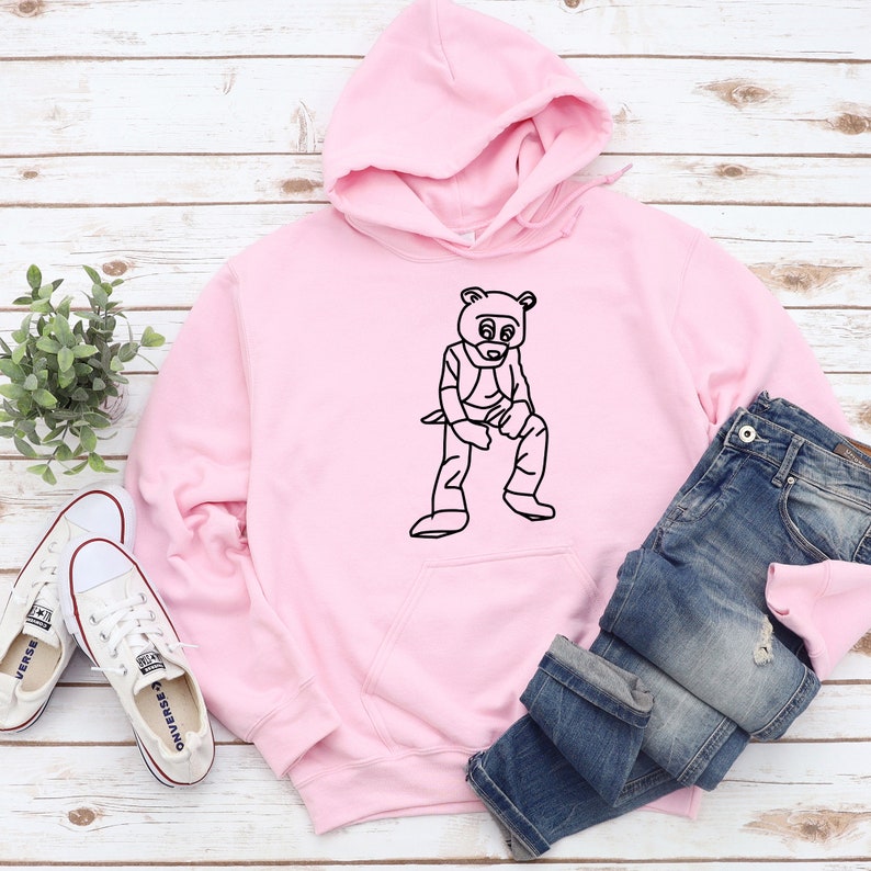 Pu&ograve; includere: Una felpa con cappuccio rosa chiaro con una grafica nera di una figura simile a un orso. La felpa &egrave; abbinata a jeans blu e scarpe da ginnastica di tela bianche. Gli articoli sono disposti su una superficie chiara con venature del legno.