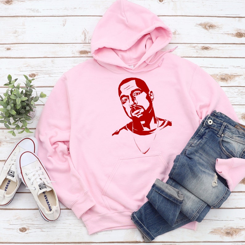 Op de afbeelding: Een lichtroze hoodie met een rode grafische afbeelding. De hoodie heeft een capuchon en een voorzak. Een blauwe spijkerbroek en witte canvas sneakers liggen naast de hoodie. De items liggen op een licht houten oppervlak.