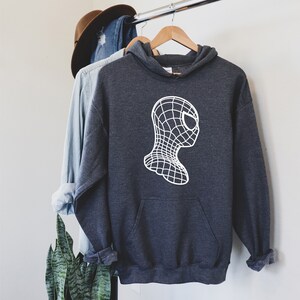 Op de afbeelding: Donkergrijze hoodie met een witte afbeelding van een gestileerd superheldenmasker. De hoodie heeft een voorzak en een capuchon met trekkoord. Andere kledingstukken en een hoed zijn zichtbaar op de achtergrond.