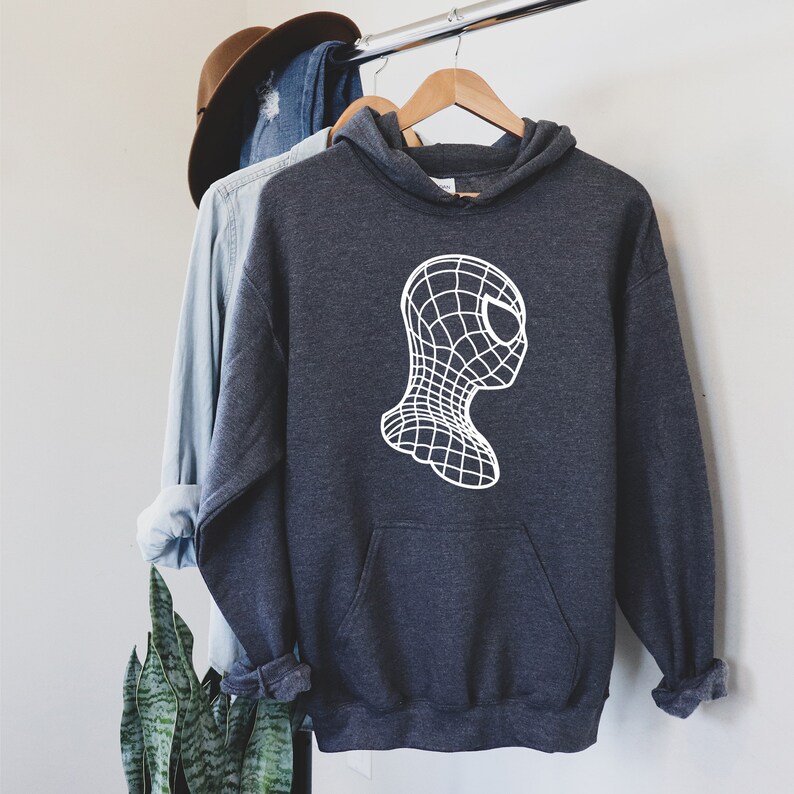 Op de afbeelding: Donkergrijze hoodie met een witte afbeelding van een gestileerd superheldenmasker. De hoodie heeft een voorzak en een capuchon met trekkoord. Andere kledingstukken en een hoed zijn zichtbaar op de achtergrond.