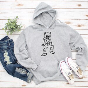 Pu&ograve; includere: Una felpa con cappuccio grigio chiaro con una grafica nera di un orso con occhiali da sole e un gilet. La felpa con cappuccio ha una tasca frontale e un cappuccio. L'immagine include anche un paio di jeans blu e scarpe da ginnastica bianche.