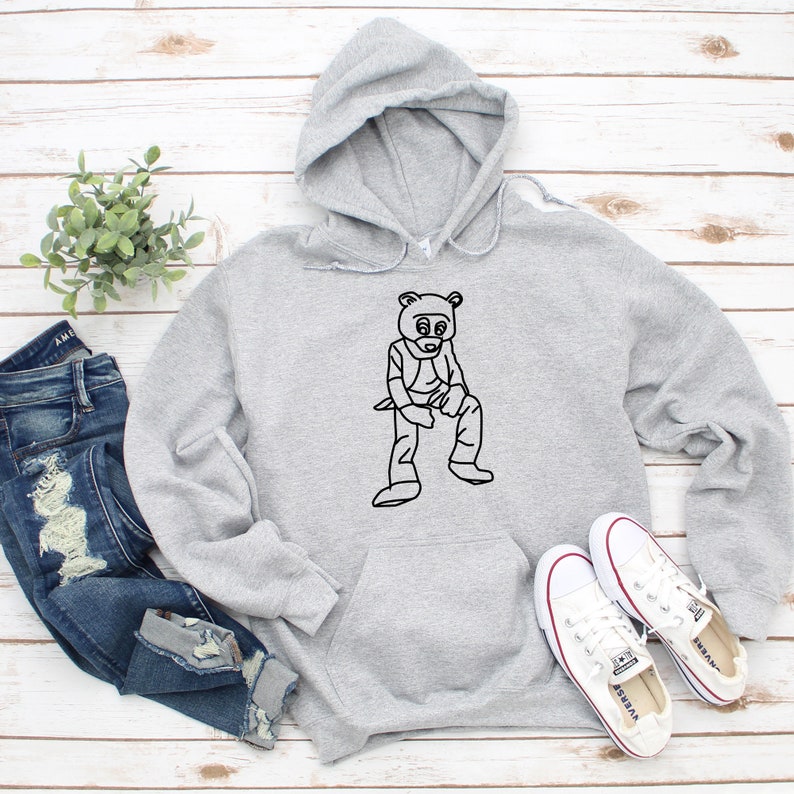 Pu&ograve; includere: Una felpa con cappuccio grigio chiaro con una grafica nera di un orso con occhiali da sole e un gilet. La felpa con cappuccio ha una tasca frontale e un cappuccio. L'immagine include anche un paio di jeans blu e scarpe da ginnastica bianche.