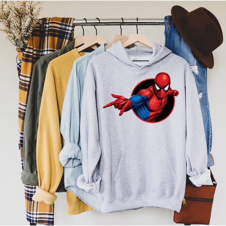 May include: A light gray hoodie featuring a graphic of Spider-Man in a black circle. The superhero is depicted in his classic red and blue suit, with his hand extended. Other clothing items, including a yellow shirt and blue jeans, are also visible.