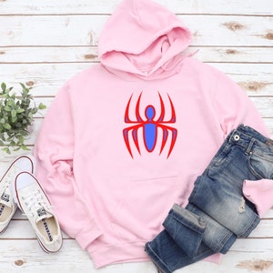 K&ouml;nnte beinhalten: Ein hellrosa Kapuzenpullover mit einem roten und blauen Spinnenmuster. Der Kapuzenpullover hat eine Kapuze, eine Vordertasche und einen Kordelzug. Eine blaue Jeans und wei&szlig;e Turnschuhe liegen neben dem Kapuzenpullover.