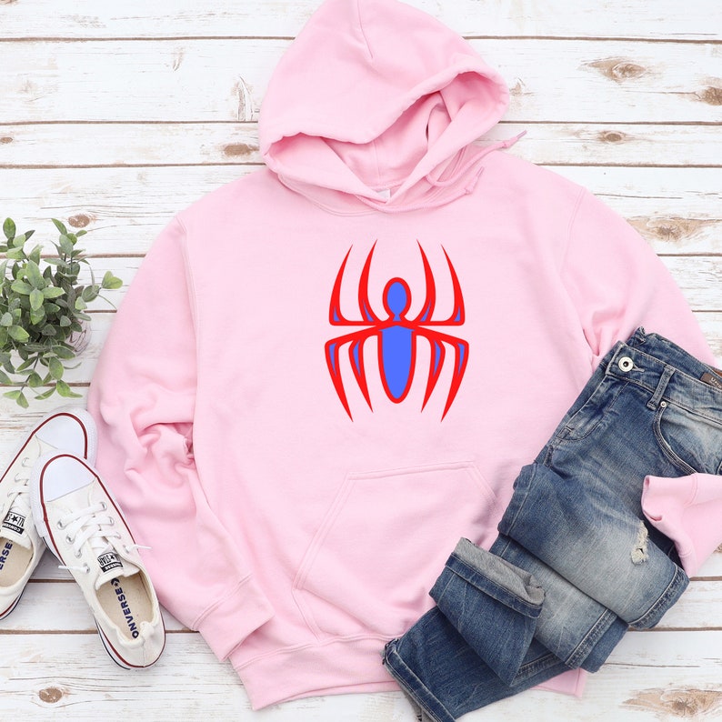 K&ouml;nnte beinhalten: Ein hellrosa Kapuzenpullover mit einem roten und blauen Spinnenmuster. Der Kapuzenpullover hat eine Kapuze, eine Vordertasche und einen Kordelzug. Eine blaue Jeans und wei&szlig;e Turnschuhe liegen neben dem Kapuzenpullover.