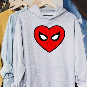 May include: A light gray hooded sweatshirt with a large red heart-shaped graphic. The graphic features a spiderweb design with white eye shapes and a black outline. The sweatshirt is hanging on a wooden hanger.