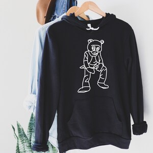 Pu&ograve; includere: Felpa con cappuccio nera con un'illustrazione bianca di un orso. La felpa ha una tasca frontale e un cappuccio con coulisse. Le maniche sono arrotolate. La felpa &egrave; appesa a un appendiabiti di legno.