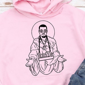 May include: A light pink hoodie featuring a black and white illustration of a person with sunglasses and a halo. The illustration includes the text "kanye" on a nameplate. A pair of blue jeans is visible in the bottom right corner.