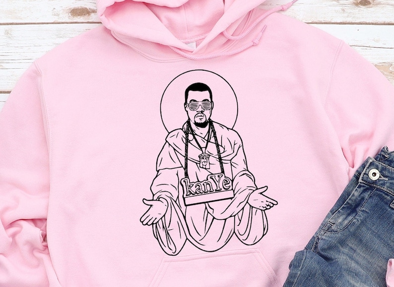 May include: A light pink hoodie featuring a black and white illustration of a person with sunglasses and a halo. The illustration includes the text "kanye" on a nameplate. A pair of blue jeans is visible in the bottom right corner.