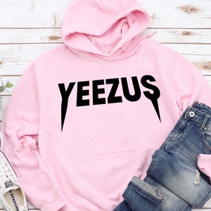 May include: A light pink hoodie with the word "YEEZUS" printed in black. The hoodie has a drawstring hood and a front pocket. Blue jeans and white sneakers are also in the image.