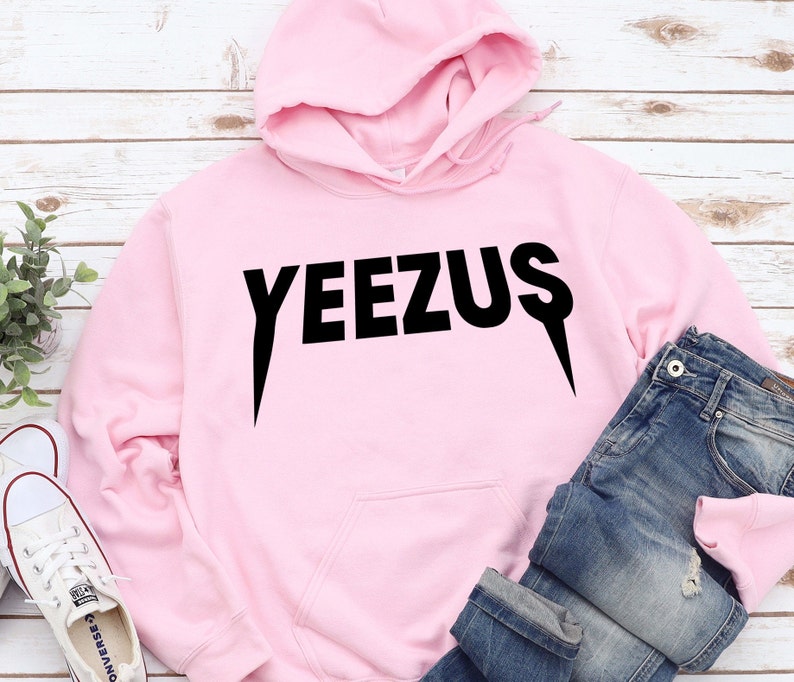 May include: A light pink hoodie with the word "YEEZUS" printed in black. The hoodie has a drawstring hood and a front pocket. Blue jeans and white sneakers are also in the image.