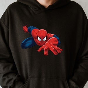 K&ouml;nnte beinhalten: Schwarzer Kapuzenpullover mit einer dynamischen Grafik von Spider-Man. Der Superheld ist in seinem klassischen rot-blauen Anzug dargestellt, mit ausgestreckten H&auml;nden. Der Kapuzenpullover ist aus einem weichen Material.