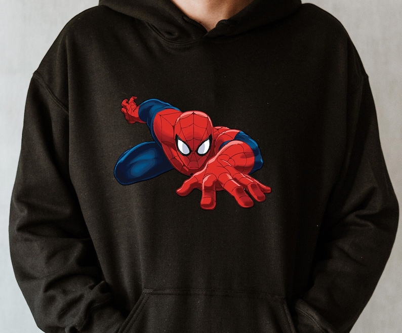 K&ouml;nnte beinhalten: Schwarzer Kapuzenpullover mit einer dynamischen Grafik von Spider-Man. Der Superheld ist in seinem klassischen rot-blauen Anzug dargestellt, mit ausgestreckten H&auml;nden. Der Kapuzenpullover ist aus einem weichen Material.