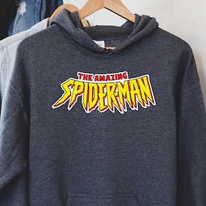 May include: A dark gray hooded sweatshirt with the words "THE AMAZING SPIDERMAN" in a yellow and red graphic. The sweatshirt is hanging on a wooden hanger.