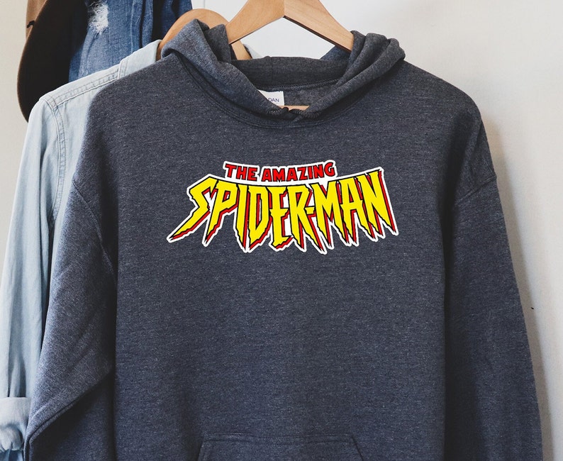 May include: A dark gray hooded sweatshirt with the words "THE AMAZING SPIDERMAN" in a yellow and red graphic. The sweatshirt is hanging on a wooden hanger.