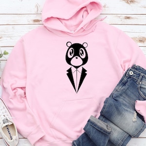 May include: A light pink hoodie featuring a black bear graphic wearing a tuxedo. The hoodie has a front pocket and a hood. Blue jeans are folded next to the hoodie. A pair of white sneakers are visible in the bottom left corner.