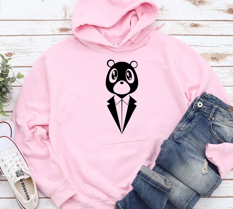 May include: A light pink hoodie featuring a black bear graphic wearing a tuxedo. The hoodie has a front pocket and a hood. Blue jeans are folded next to the hoodie. A pair of white sneakers are visible in the bottom left corner.