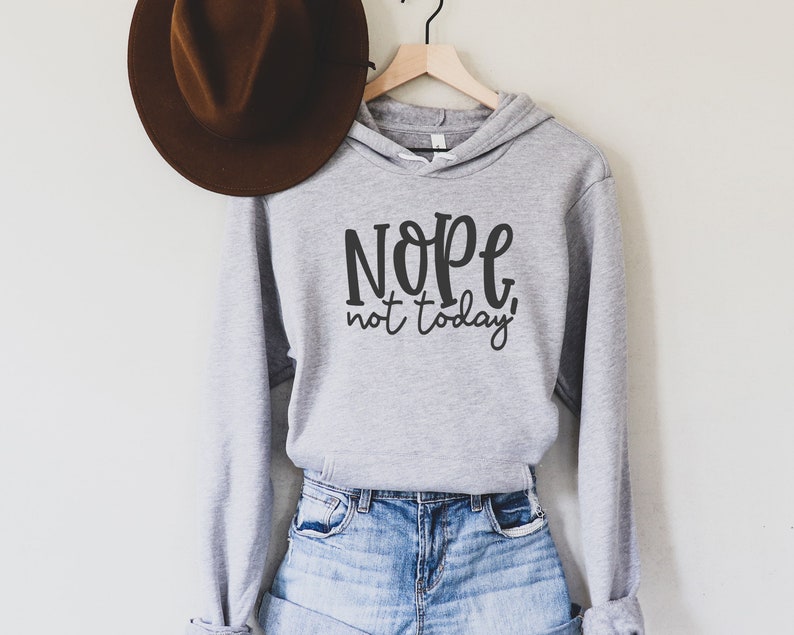 May include: A pale grey hoodie with the text "Nope, not today!" in black script. The hoodie is paired with denim shorts. A brown hat hangs on a hook above the hoodie.