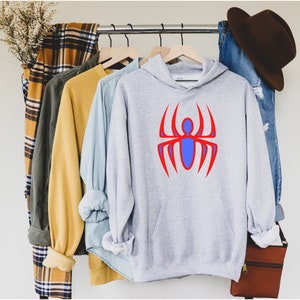 K&ouml;nnte beinhalten: Ein hellgrauer Kapuzenpullover mit einem roten und blauen Spinnenmuster. Der Kapuzenpullover h&auml;ngt zusammen mit anderen Hemden und einer Jeans an einer Kleiderstange. Ein brauner Hut und eine Tasche sind ebenfalls zu sehen.