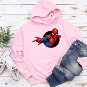 May include: A light pink hoodie featuring a Spider-Man graphic. The graphic shows Spider-Man in his classic red and blue suit, with a black circle background. The hoodie is paired with blue jeans and white sneakers.