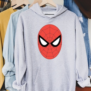May include: A light gray hooded sweatshirt featuring a large red Spider-Man mask design on the front. The mask has a black outline, white eye shapes, and a web pattern. The sweatshirt is hanging on a wooden hanger.