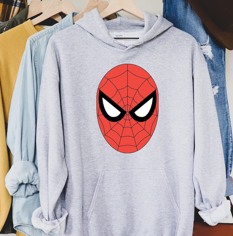May include: A light gray hooded sweatshirt featuring a large red Spider-Man mask design on the front. The mask has a black outline, white eye shapes, and a web pattern. The sweatshirt is hanging on a wooden hanger.