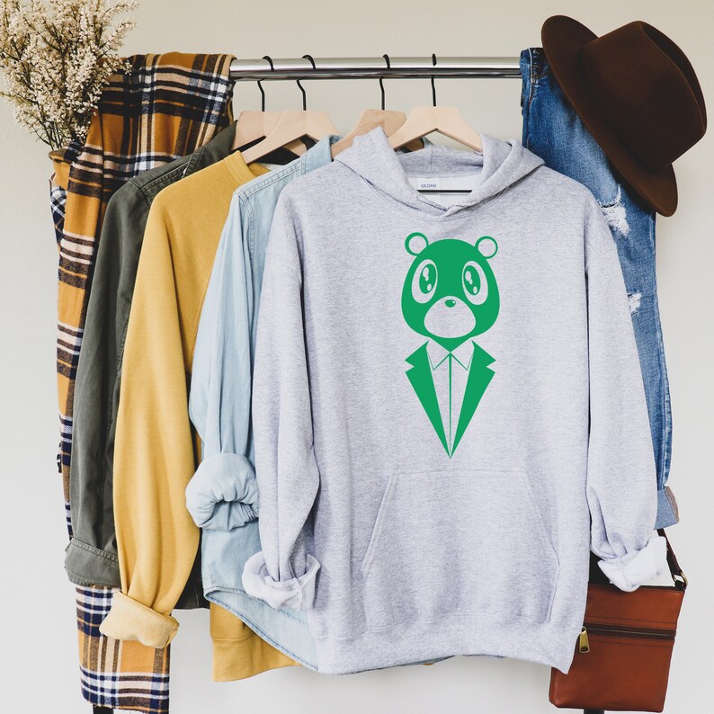 May include: A light gray hoodie with a green bear graphic wearing a suit. The hoodie is hanging on a wooden hanger with other clothing items, including a yellow shirt, a denim shirt, and a plaid scarf. A brown hat and jeans are also visible.