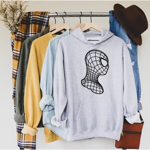 Op de afbeelding: Een lichtgrijze hoodie met een zwarte afbeelding van een gestileerde Spider-Man-kop. De hoodie heeft een voorzak en hangt aan een kledingrek met andere shirts en een spijkerbroek. Een bruine hoed en tas zijn ook zichtbaar.