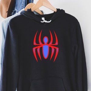 K&ouml;nnte beinhalten: Schwarzer Kapuzenpullover mit roter und blauer Spinnengrafik. Das Spinnendesign befindet sich mittig auf der Vorderseite des Sweatshirts. Das Sweatshirt h&auml;ngt an einem Holzkleiderb&uuml;gel.