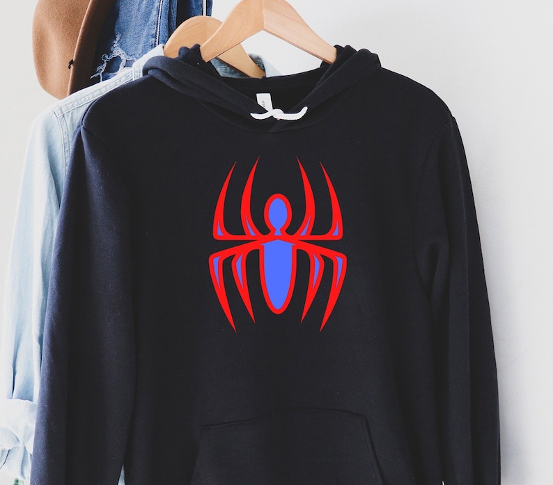 K&ouml;nnte beinhalten: Schwarzer Kapuzenpullover mit roter und blauer Spinnengrafik. Das Spinnendesign befindet sich mittig auf der Vorderseite des Sweatshirts. Das Sweatshirt h&auml;ngt an einem Holzkleiderb&uuml;gel.