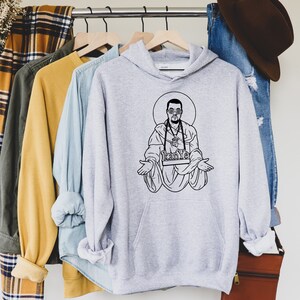 May include: A light gray hoodie with a black graphic of a man in sunglasses, a halo, and the word "Kanye" on a banner. The hoodie is hanging on a wooden hanger with other clothing items.