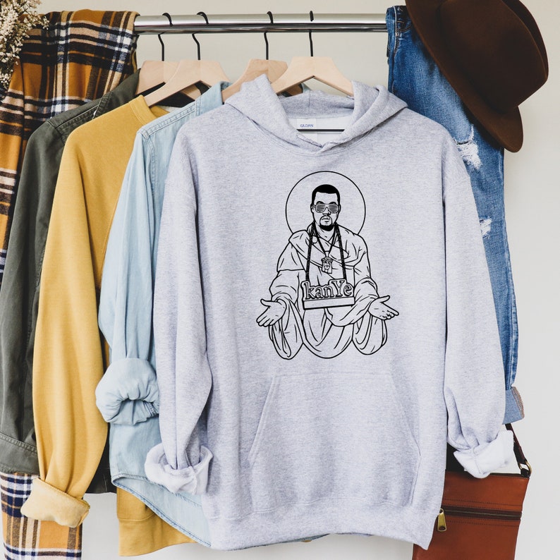 May include: A light gray hoodie with a black graphic of a man in sunglasses, a halo, and the word "Kanye" on a banner. The hoodie is hanging on a wooden hanger with other clothing items.