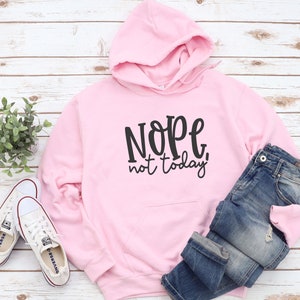 May include: A pale pink hoodie with the phrase "Nope, not today!" printed in black. The hoodie is displayed with blue denim jeans and white trainers. The items are arranged on a light wood-effect surface.