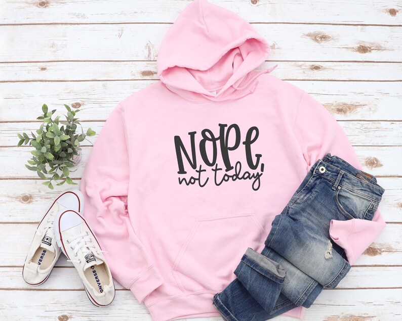 May include: A pale pink hoodie with the phrase "Nope, not today!" printed in black. The hoodie is displayed with blue denim jeans and white trainers. The items are arranged on a light wood-effect surface.